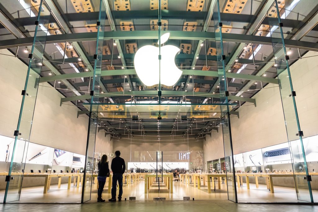 Modern Apple Store with large glass entrance and illuminated Apple logo, showcasing minimalist interior design and customers browsing technology products.