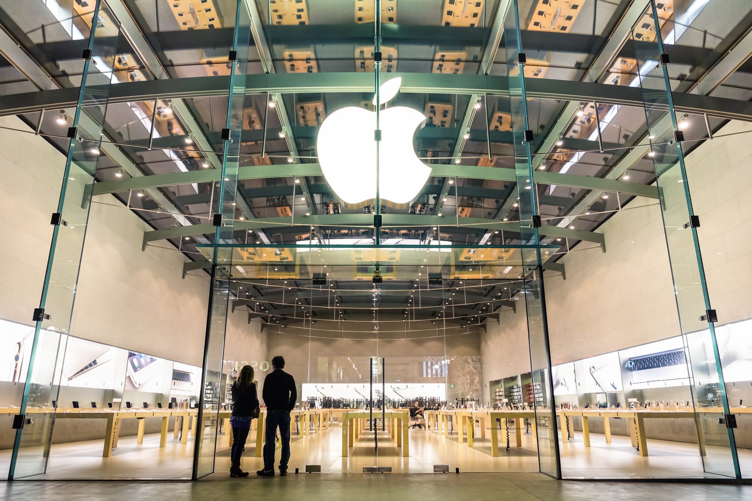 Modern Apple Store with large glass entrance and illuminated Apple logo, showcasing minimalist interior design and customers browsing technology products.