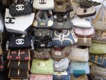 Various fake designer handbags displayed on a market stall, including styles with quilted patterns and logos resembling luxury brands.