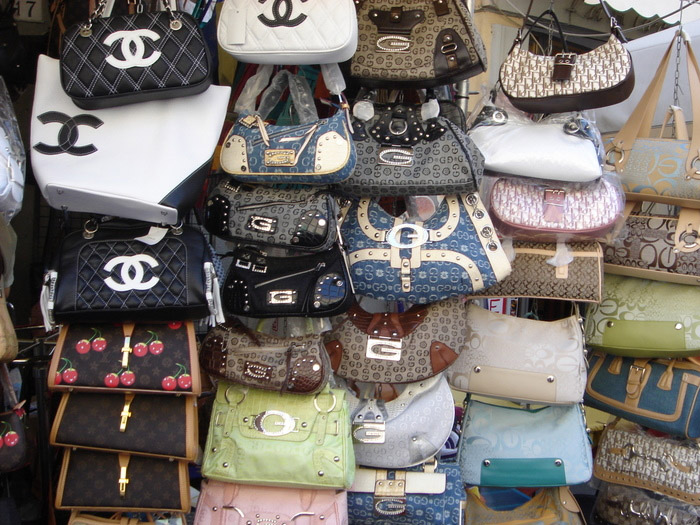 Various fake designer handbags displayed on a market stall, including styles with quilted patterns and logos resembling luxury brands.