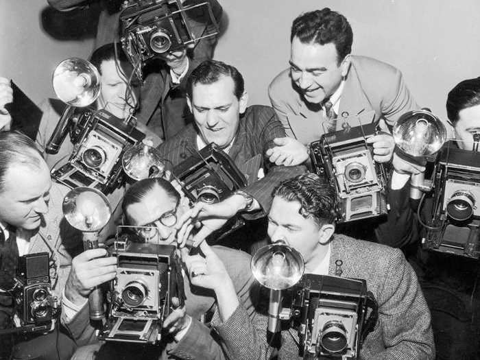 Group of vintage photographers using old-fashioned cameras with flashbulbs, capturing a photo in a crowded setting.